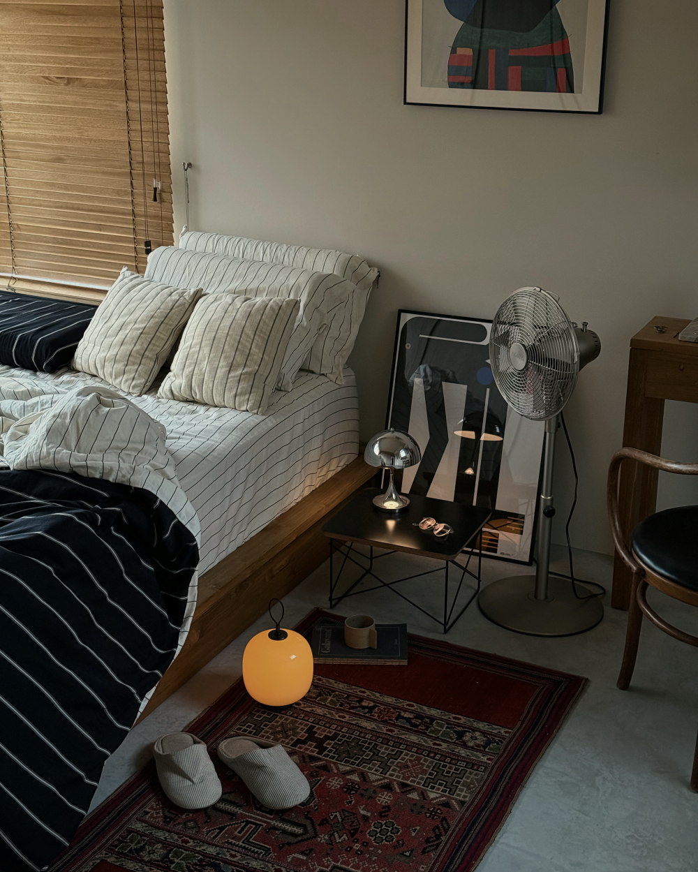 A glowing VL 45 Radiohus Portable Lamp on the floor by a bed with striped bedding and a rug, © 2025 Louis Poulsen.