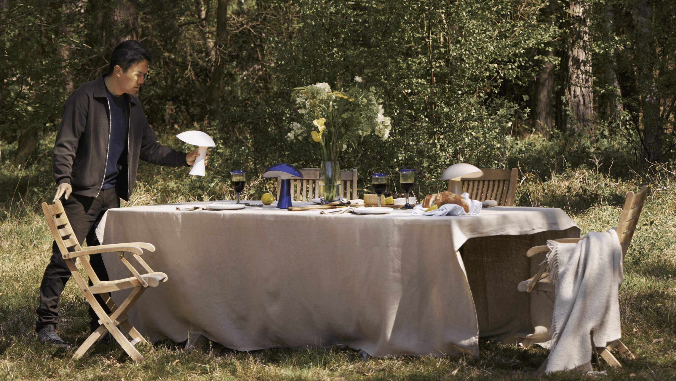 Gabriel Tan setting the Rumee portable lamp on a table during an outdoor gathering, with a natural backdrop, © 2025 Louis Poulsen.